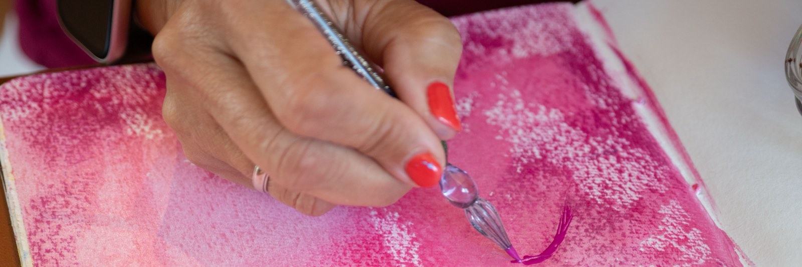 Annett schreibt mit einer gläsernen Feder in pinker Tinte auf eine rosa bemalte Seite in ihrem Skizzenbuch.