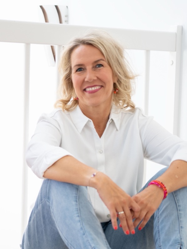 Annett Liskewitsch sitzt auf dem Boden vor einer hellen Wand, die Arme locker auf den Knien, und lächelt in die Kamera.