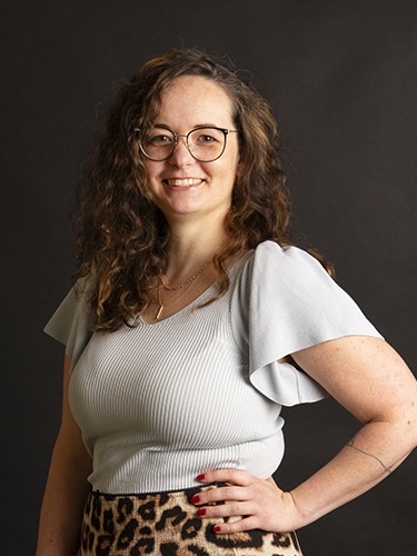 Sarah Hübner steht vor dunklem Hintergrund, eine Hand in der Hüfte, und lächelt in die Kamera.